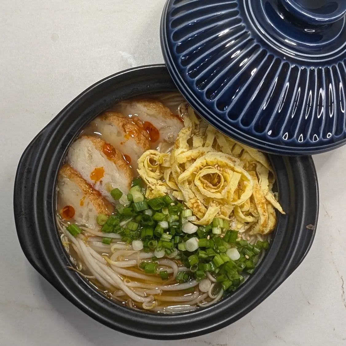 Takana Tonkotsu ramen in a bowl topped with sliced golden onion fish cakes, Kinshi Tamago egg ribbons, bean sprouts, green onions, toasted sesame seeds, and chili oil.